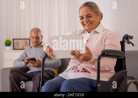 Donna anziana che usa il tablet digitale su sedia a rotelle e uomo anziano che usa lo smartphone sul divano Foto Stock