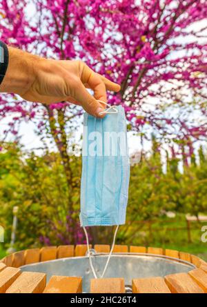 Maschi uomo dita tiene la maschera blu viso sopra il bidone con la natura primavera in fiore sullo sfondo. Concetto di smaltimento delle maschere e fine della quarantena e. Foto Stock