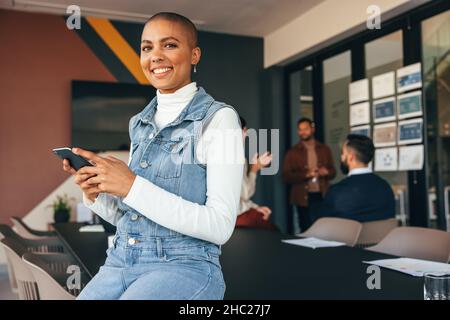 Donna d'affari millenaria che sorride alla fotocamera mentre tiene uno smartphone. Giovane donna d'affari allegra seduta su un tavolo della sala riunioni con il suo collegu Foto Stock