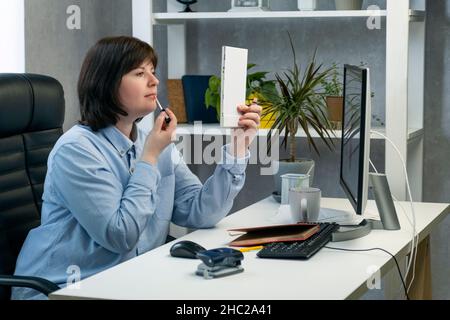 Donna che fa trucco e preening sul posto di lavoro. Ragazza dipinge le labbra al lavoro in ufficio privato Foto Stock