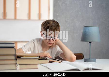 Scolaro in occhiali fa i suoi compiti. Apprendimento a distanza. Ragazzo legge il libro di testo. Homeschooling. Foto Stock