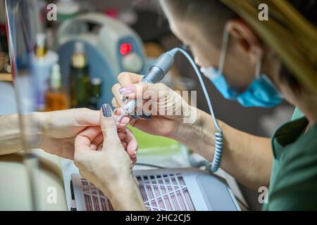 Donna nel salone delle unghie che riceve manicure nel salone di bellezza da estetista che usano il trapano elettrico della lima delle unghie con la polvere di volo di scoppio e detriti intorno. Concetto di cura delle unghie. Foto Stock