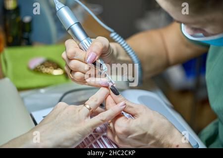 Donna nel salone delle unghie che riceve manicure nel salone di bellezza da estetista che usano il trapano elettrico della lima delle unghie con la polvere di volo di scoppio e detriti intorno. Concetto di cura delle unghie. Foto Stock