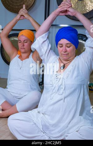 Due donne vestite di bianco con il turbante che praticano lo yoga kundalini seduto sul pavimento con le mani sulla testa Foto Stock