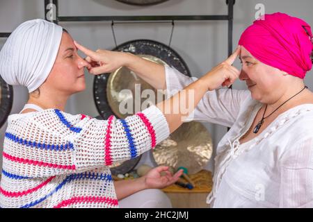 Due donne vestite di bianco con turbante che fanno kundalini yoga seduti sul pavimento con il dito indice sulla fronte l'una dell'altra Foto Stock