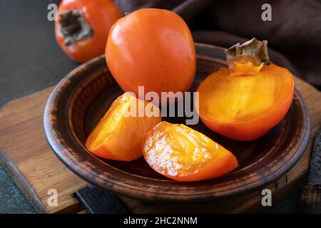 Frutta fresca biologica matura Persimmons in ciotola di legno con fetta su sfondo di un tavolo di pietra scura. Cibo da dessert, cibo vegano sano. Foto Stock