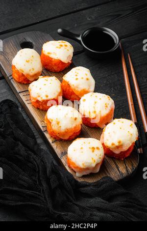 Panini maki al forno con salmone, granchio, cetriolo, avocado, roe di pesce volante e salsa piccante, su sfondo nero tavola di legno Foto Stock