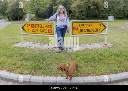 Turista maturo sorridente e guardando la macchina fotografica, insieme al suo cane marrone in piedi tra un segnale di direzione informativo a un bivio, un paese ro Foto Stock