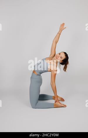 La brunetta si pone su sfondo isolato. Giovane donna in tuta grigia per il fitness o lo yoga fa esercizi stretching, ginocchia e solleva la mano, lato Foto Stock