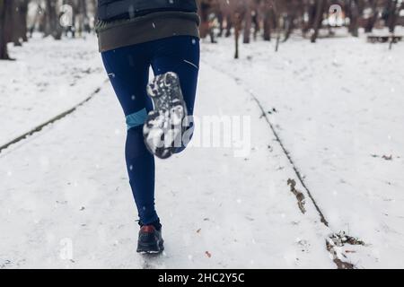 Atleta giovane donna che corre nel nevoso parco invernale. Primo piano di sneakers e gambe. Stile di vita sano e attivo Foto Stock