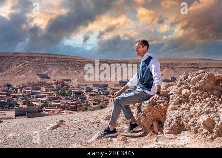 Giovane uomo seduto su roccia contro la vista panoramica della terra deserta e cielo nuvoloso Foto Stock