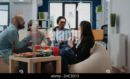 I colleghi che giocano a charades gioco con note adesive dopo il lavoro in ufficio. I compagni di lavoro si divertiranno a indovinare il gioco per fare attività e divertimento mentre bevono birra e mangiano pizza. Foto Stock