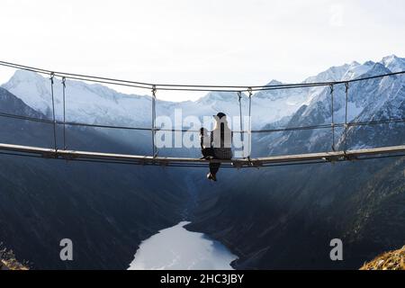 Ragazza viaggiatore con il suo cane in montagna. Una ragazza siede su un ponte sospeso. Foto Stock