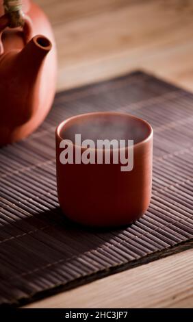 Tazza di tè o caffè con vapore caldo di argilla marrone, su tavola di legno con vista parziale della teiera o della caffettiera. Sfondo marrone. Foto Stock