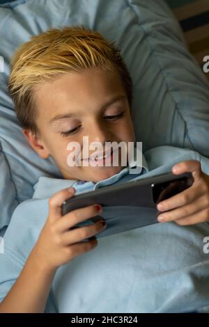 Il bambino è immerso in un abbigliamento casual sdraiato sul letto e giocare a videogiochi sul telefono cellulare mentre si passa del tempo libero a casa Foto Stock