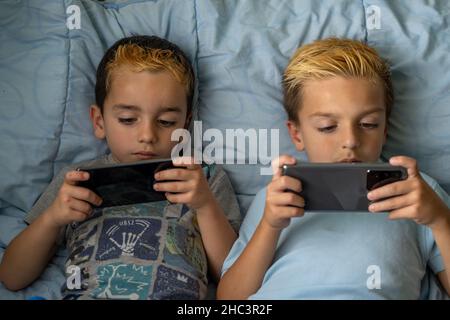 Da sopra dei fratelli freschi di preschool con i capelli tinti che si trovano insieme sul letto e sulla navigazione degli smartphone mentre trascorrono il tempo libero a casa Foto Stock