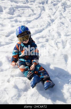 il bambino stanco in un casco, occhiali, scarponi da sci e tute invernali si siede nella neve in una stazione sciistica. Lezione di sci per bambini in una scuola alpina. Caduto dentro Foto Stock