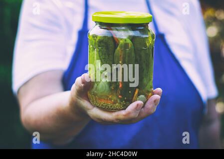 Donna anziana che tiene un vaso di sottaceti nel giardino. Cetrioli sott'aceto fatti in casa in un vasetto di vetro. Vasetto di conservazione di cetriolini con semi di senape e aneto Foto Stock