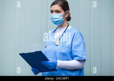 Ritratto di giovane bella caucasica femmina medico medico medico NHS medico, in piedi di fronte a parete di vetro blu, guardando via, indossare l'attrezzatura di protezione blu, fa Foto Stock