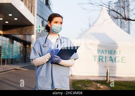 Giovane bella caucasica femmina NHS medico di fronte alla clinica di ambulanza di emergenza, accanto al paziente bianco ammissione tenda di triage per ricevere infetto Foto Stock