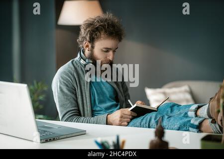 Lo studente ascolta una lezione online e prende appunti in un notebook. Un uomo scrive informazioni importanti in un diario. Cuffie intorno al collo. Piedi sul tavolo. Primo piano. Foto di alta qualità Foto Stock