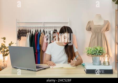 Donna asiatica imprenditore di negozio di abbigliamento online di lavoro e prendere nota in notebook con notebook sulla scrivania in ufficio a casa Foto Stock