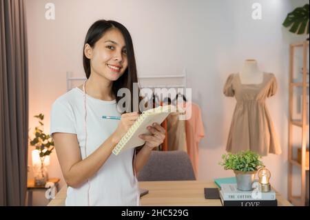 Donna asiatica felice imprenditore in piedi e prendendo nota in negozio di abbigliamento online Foto Stock