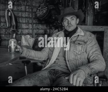 Scatto in scala di grigi di un cowboy seduto su una sedia che beve birra Foto Stock