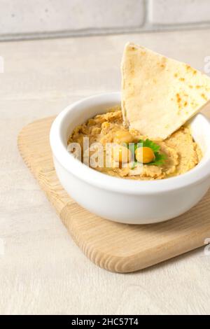 Hummus in una ciotola bianca di ceramica con pezzi di pita su un asse di legno su uno sfondo chiaro. Un piatto tradizionale mediorientale. Orientamento orizzontale. Foto Stock