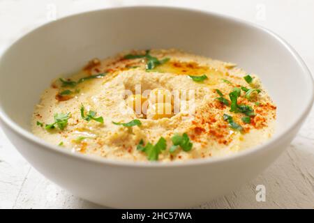 Hummus in un recipiente bianco in ceramica con foglie di paprika e prezzemolo su sfondo chiaro. Un piatto tradizionale mediorientale. Orientamento orizzontale. Veget Foto Stock