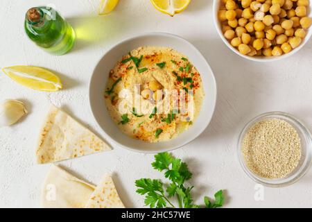 Hummus in un recipiente bianco in ceramica con foglie di paprika e prezzemolo e ingredienti su sfondo chiaro. Un piatto tradizionale mediorientale. Orizzontale o. Foto Stock