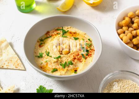 Hummus in un recipiente bianco in ceramica con foglie di paprika e prezzemolo e ingredienti su sfondo chiaro. Un piatto tradizionale mediorientale. Orizzontale o. Foto Stock