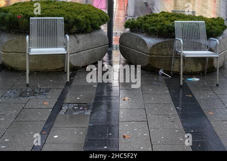 Due sedie pubbliche di metallo di fronte a due grandi vasi di pianta di pietra su pioggia imbevuto terreno sporco su una piazza. Foto Stock