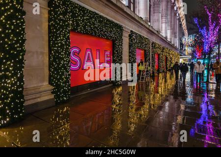 Gli acquirenti fuori Selfridges in una serata piovosa come i cartelli di vendita sono messi in su la vigilia di Natale a Londra. Foto Stock