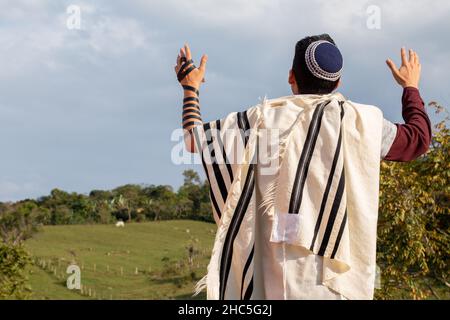 Foto orizzontale di Juido Latino indossando tefilin e kippah alzando le mani pregando guardando il cielo con uno sfondo bellissimo di natuleza Foto Stock