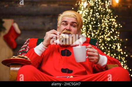 Babbo Natale mangiare biscotti e bere latte. Uomo bearded in costume di Santa che si rilassa a casa. Natale. Foto Stock