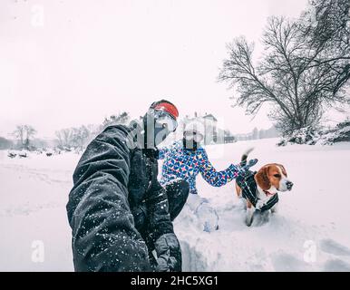 Coppia felice con il loro cane beagle, prendendo un selfie durante una tempesta di neve, dotato di abbigliamento invernale, closeup, concetto di vacanza Foto Stock
