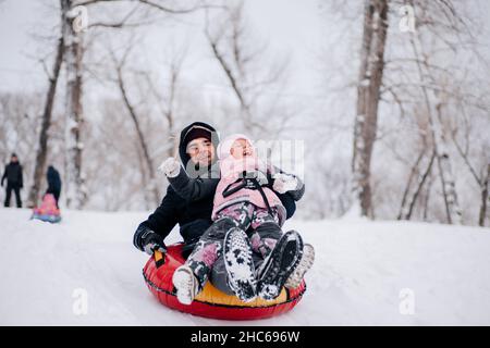 il ragazzino sorride e scivola giù per la collina sulla slitta con il padre che indossa abiti invernali caldi nella foresta. Sfondo sorprendente pieno di colore bianco e. Foto Stock