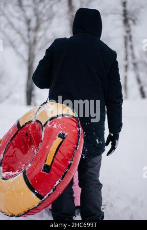 Primo piano foto di femmina russo capretto non visto dietro il padre con slitta che va su collina per scivolare giù nella neve nella foresta. Sfondo sorprendente pieno di bianco Foto Stock