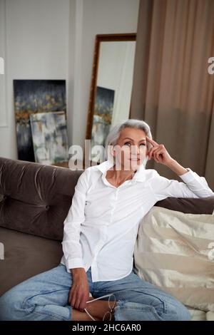 Alto angolo di affascinante femmina di mezza età con capelli grigi e in jeans e camicia bianca seduta con gambe incrociate sul divano a casa e sorridente alla macchina fotografica Foto Stock