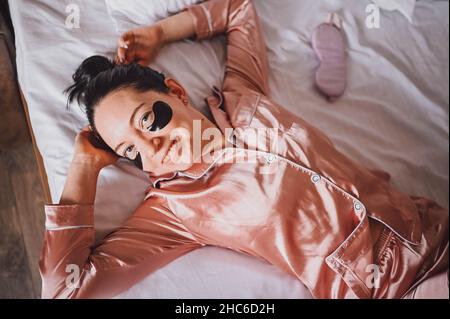 Cosmetologia, cura della pelle, trattamento del viso, spa, concetto di bellezza naturale. Giovane donna sorridente in pigiama rosa di seta si trova a letto con macchie nere idratanti sugli occhi. Routine di bellezza Foto Stock