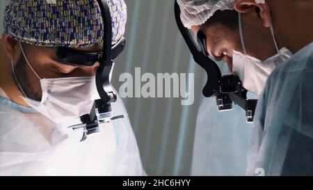 Team medico che esegue l'operazione. Primo piano dei volti dei chirurghi al lavoro indossando le feritoie binoculari in sala operatoria durante la procedura. Foto Stock