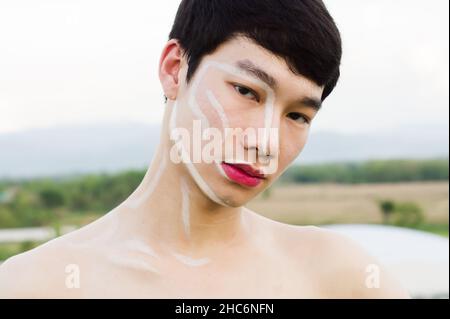 Un uomo gay asiatico bello che non indossa una camicia, scrive sul viso e indossa un rossetto rosa. Spazio di copia Foto Stock