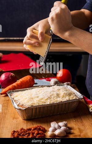 Immagine verticale di una persona irriconoscibile che grattugiava formaggio fresco sulle lasagne con una grattugia con diversi tipi di ingredienti intorno al cibo. Foto Stock