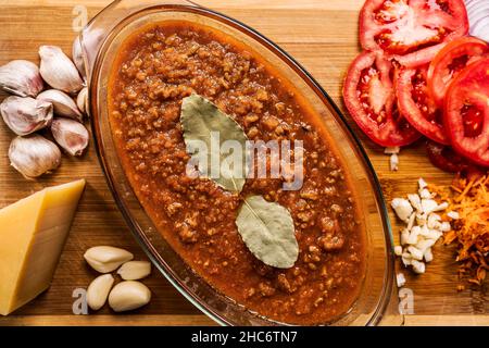 Vista dall'alto di una salsa di bolognese fatta in casa con foglie di alloro in una grande ciotola di vetro con diversi tipi di verdure intorno come fette di pomodori, aglio Foto Stock