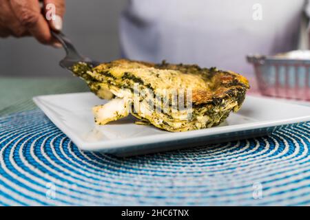 Vista ravvicinata di una mano di una donna adulta che serve una porzione di lasagna pesto appena fatta su un piatto. Foto Stock