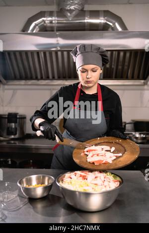 Donna chef in abiti speciali e guanti di gomma, versando bastoncini di granchio a fette in una ciotola. Concetto di preparazione dell'insalata dietetica. Foto Stock