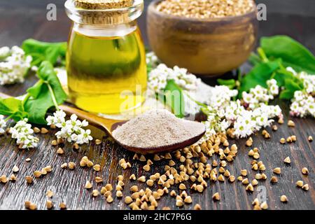 Farina di grano saraceno da cereali marroni in un cucchiaio, olio in un vaso di vetro su insaccamento, fiori e foglie di grano saraceno su sfondo legno asse Foto Stock