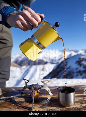 Uomo escursionista versare il caffè da giallo Moka mocha pot all'aperto nella neve inverno montagne. Vecchio stile caffè vintage pot campeggio all'aperto Foto Stock