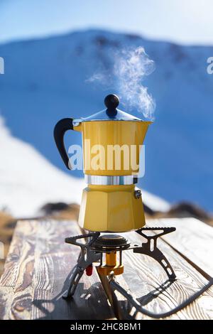 Caffè giallo moka pot con vapore al campeggio neve montagna. Picnic mattutino, persona che cucinano una bevanda calda per colazione nella natura Foto Stock